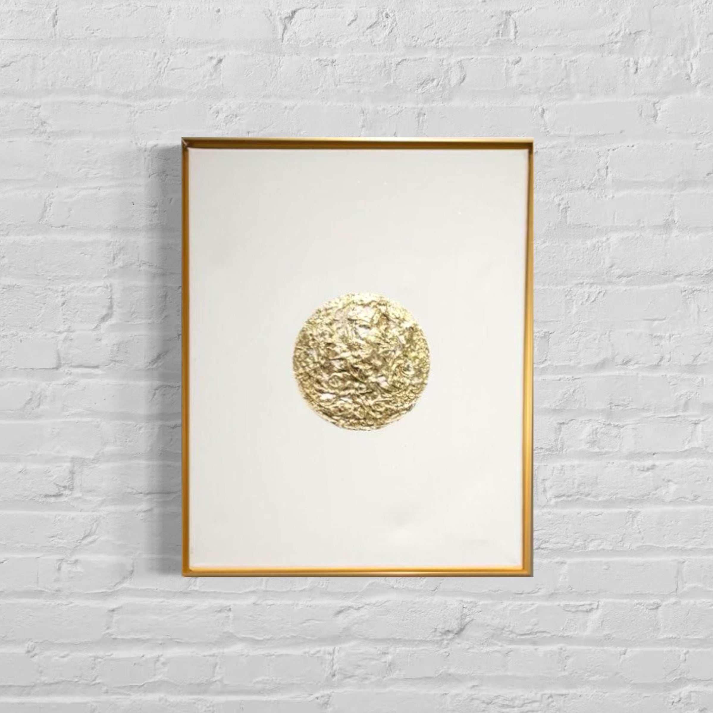 Framed artwork with a gold circular design on a white background, mounted on a light gray brick wall.